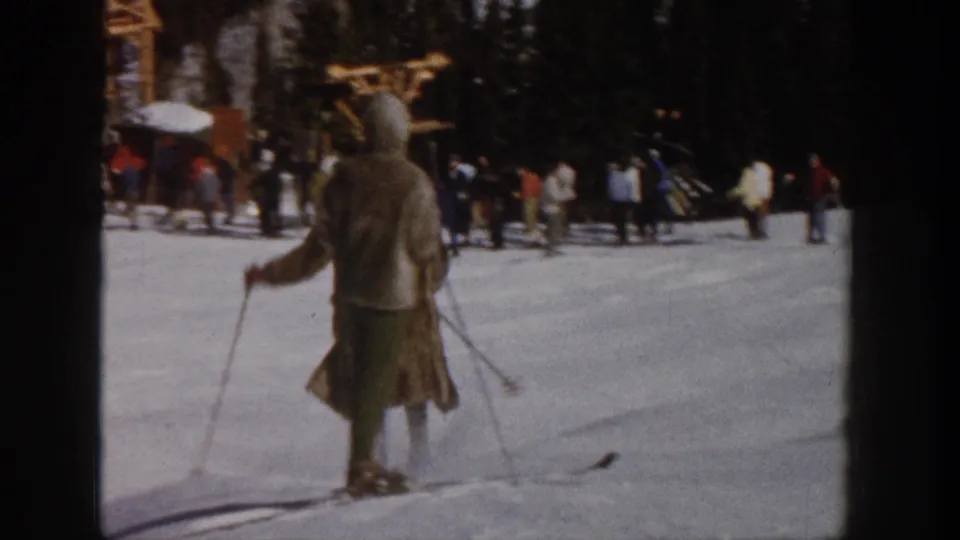 1961 Aspen frame showing an adult skier from behind near spectators, trees, and a lodge-like structure at the edge of the snow.