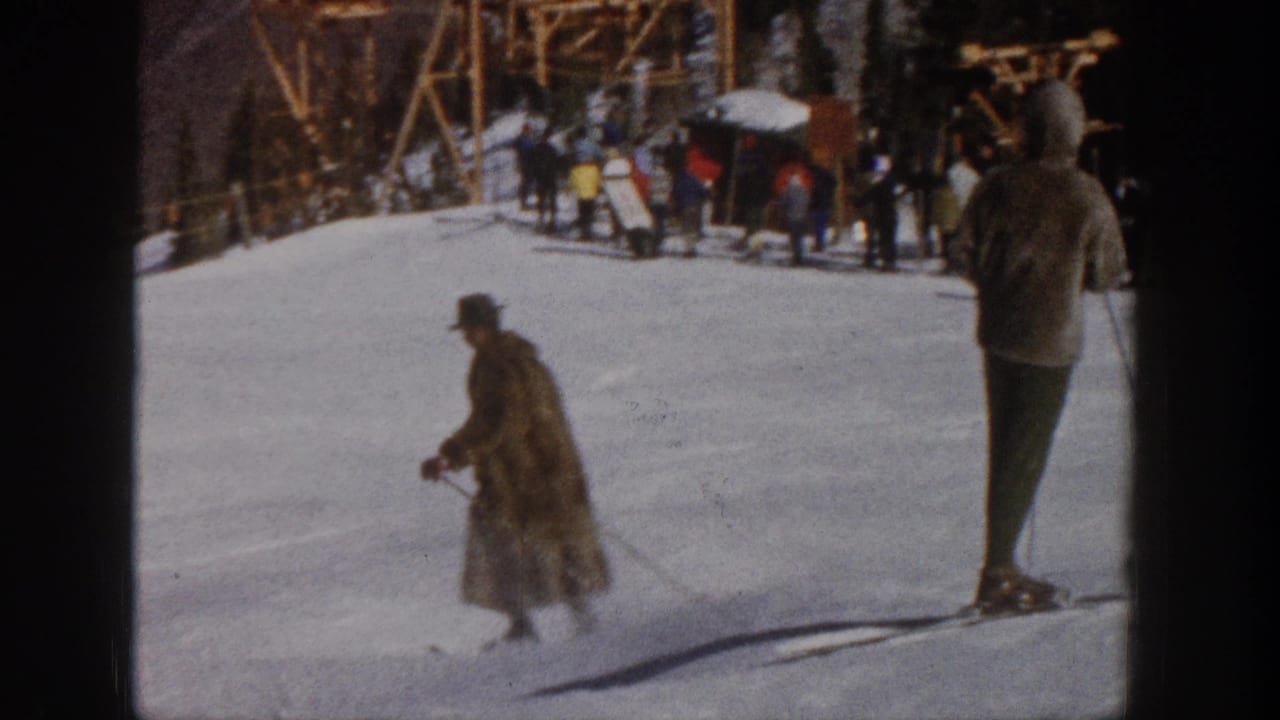 1961 Aspen home-movie frame showing a coated man skiing downhill while other skiers gather near a wooden slope-side structure.