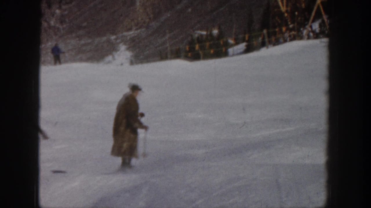 1961 Aspen frame with a man in a long coat and hat skiing across a pale snowy run, backed by trees and a distant fence line.