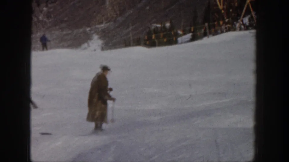 1961 Aspen frame with a man in a long coat and hat skiing across a pale snowy run, backed by trees and a distant fence line.