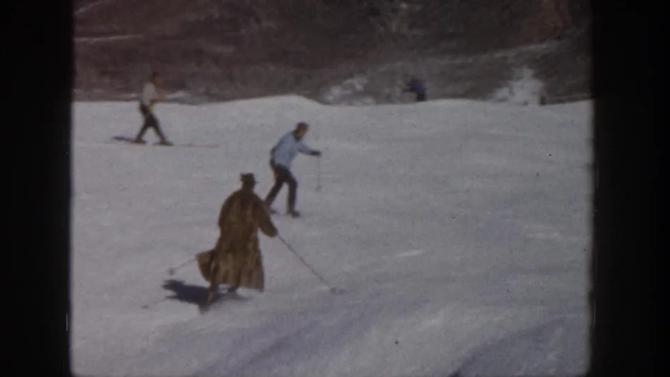 1961 Aspen home-movie still showing the coated skier from behind on a snowy run, with other skiers spaced across the slope.