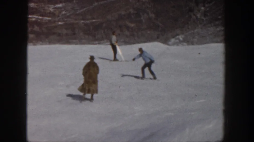 1961 Aspen frame showing the coated skier lower on the slope, with two other skiers nearby against a wooded winter background.