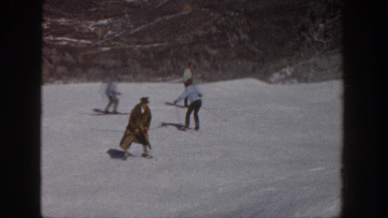 1961 Aspen home-movie frame showing skiers on a snowy slope, with one long-coated skier appearing off balance in the foreground.