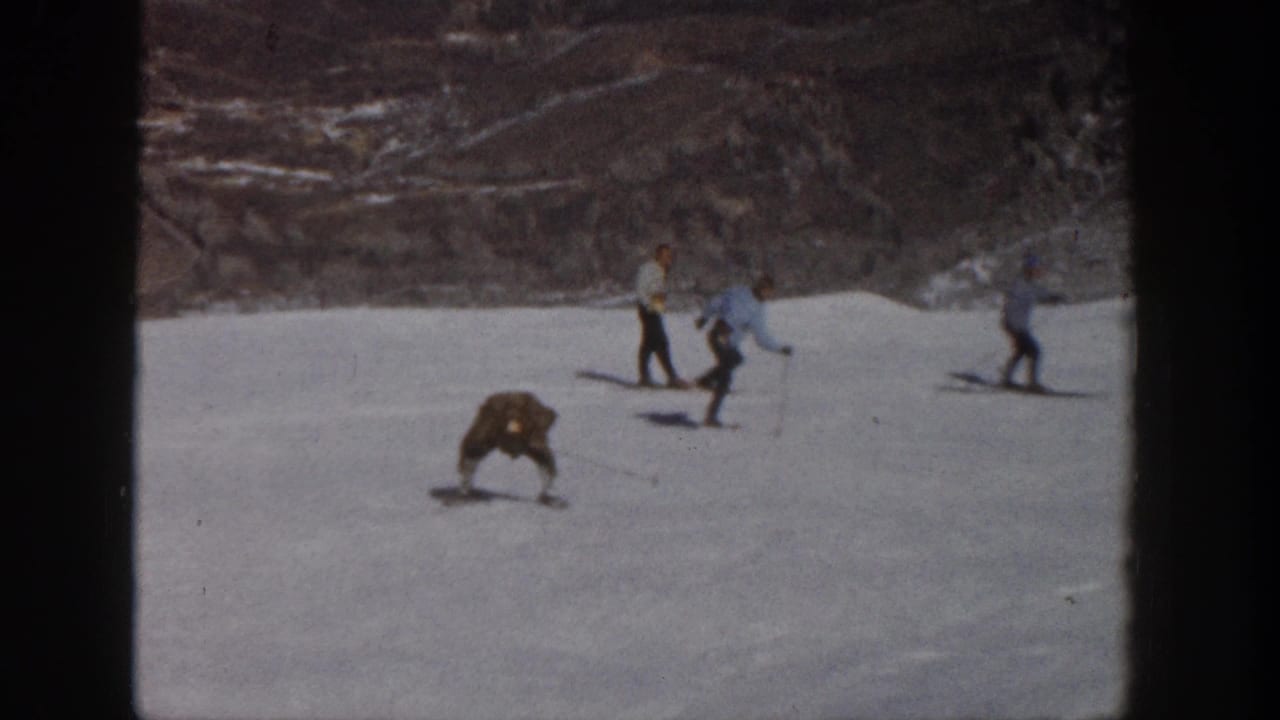 1961 Aspen ski-slope frame with a long-coated skier bent low as nearby skiers move across the snowy background.