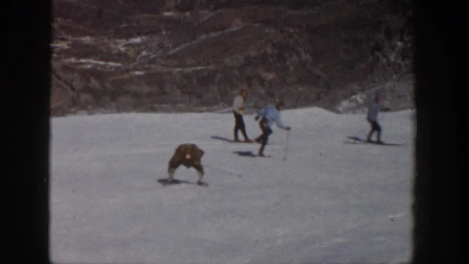 1961 Aspen ski-slope frame with a long-coated skier bent low as nearby skiers move across the snowy background.