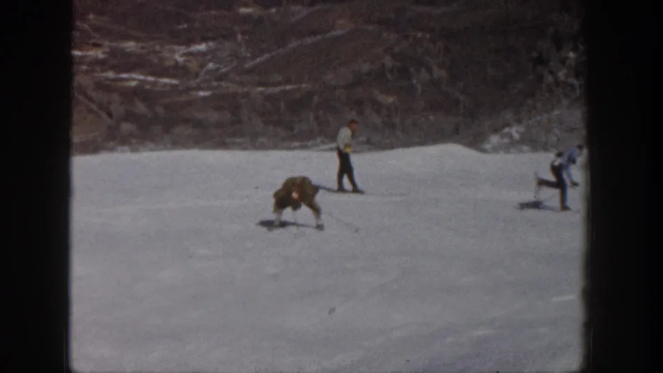 1961 Aspen home-movie frame showing the long-coated skier down on the snowy slope with another skier standing nearby.