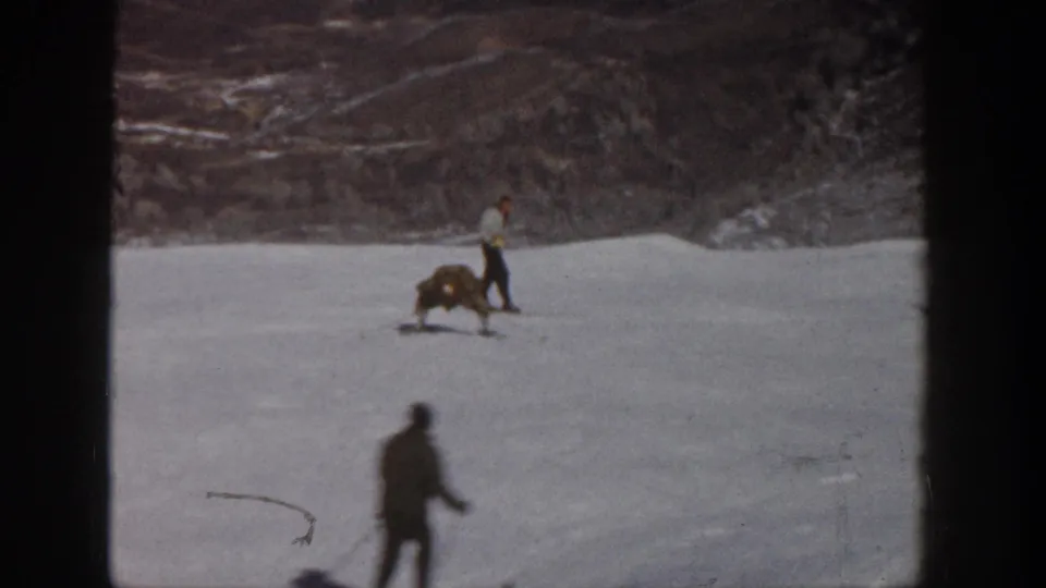 1961 Aspen ski-slope frame with the fallen skier in midground and a foreground skier moving past on snow.