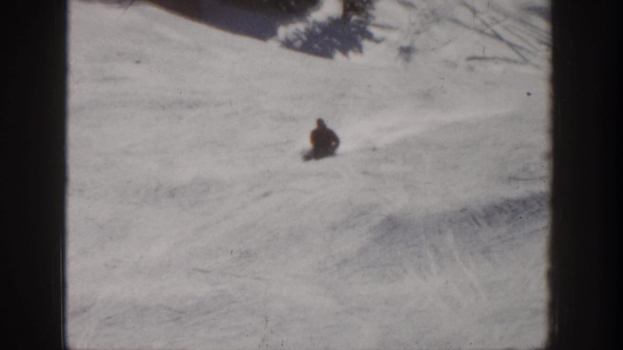 A distant skier appears low to the snow on an Aspen ski slope, with broad tracked snow around them.