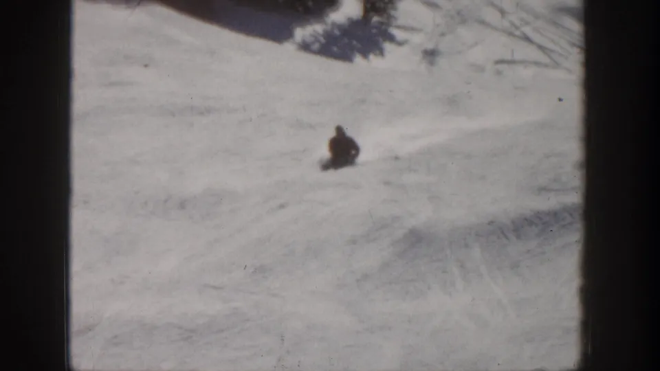 A distant skier appears low to the snow on an Aspen ski slope, with broad tracked snow around them.