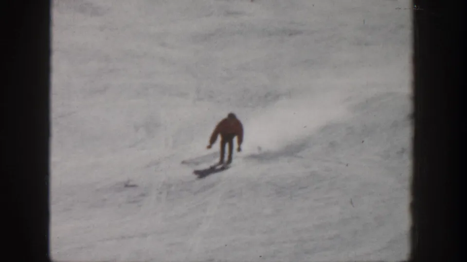 1961 Aspen frame showing a skier in a red jacket leaning forward while attempting to descend a snowy slope.