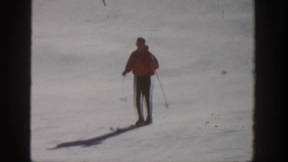 A skier in a red jacket stands on skis with poles on a sunlit Aspen slope in 1961 home-movie footage.