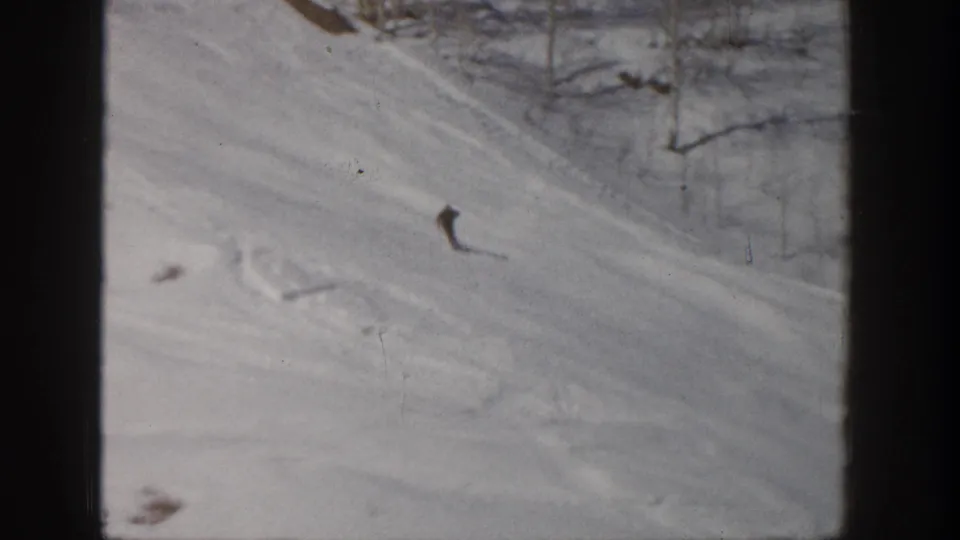 1961 Aspen home-movie frame showing a distant skier turning down a snow-covered mountain slope bordered by bare trees.