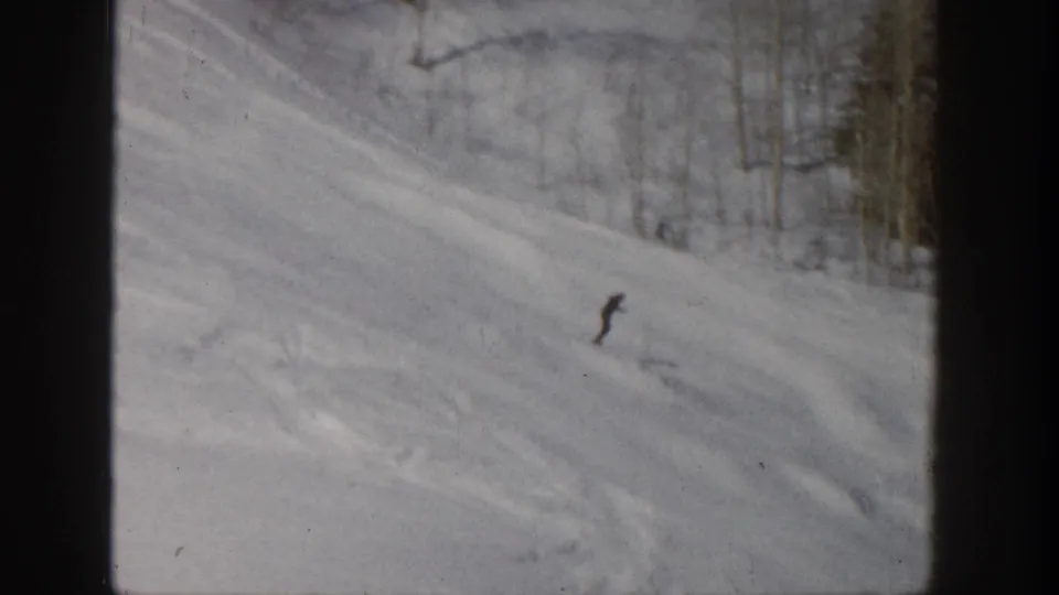 1961 Aspen 8mm frame showing the skier farther downhill on a broad snowy slope with winter trees behind.
