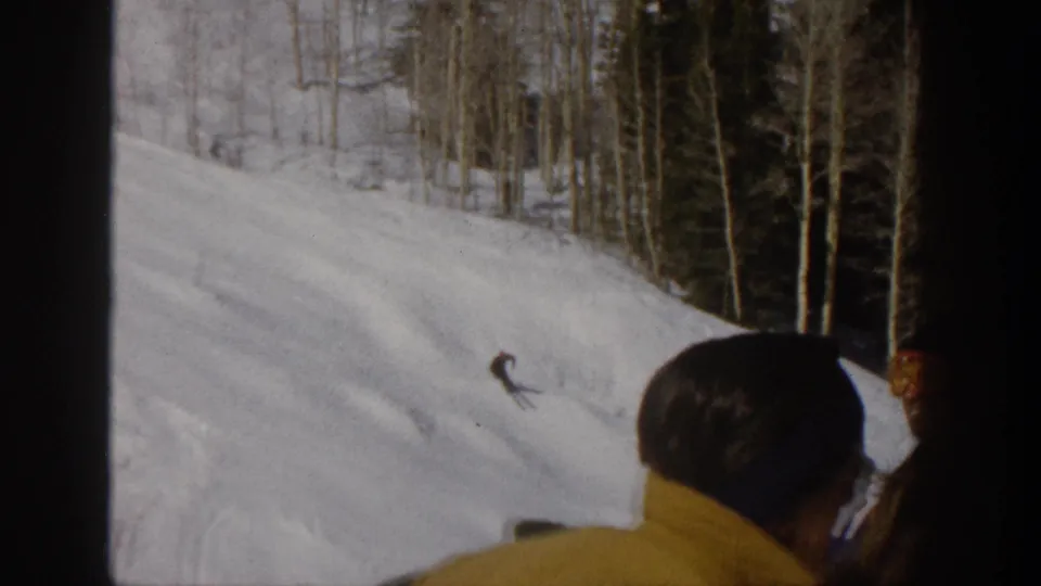 1961 Aspen frame with two onlookers in the foreground watching a distant skier on a snowy, tree-lined mountain slope.