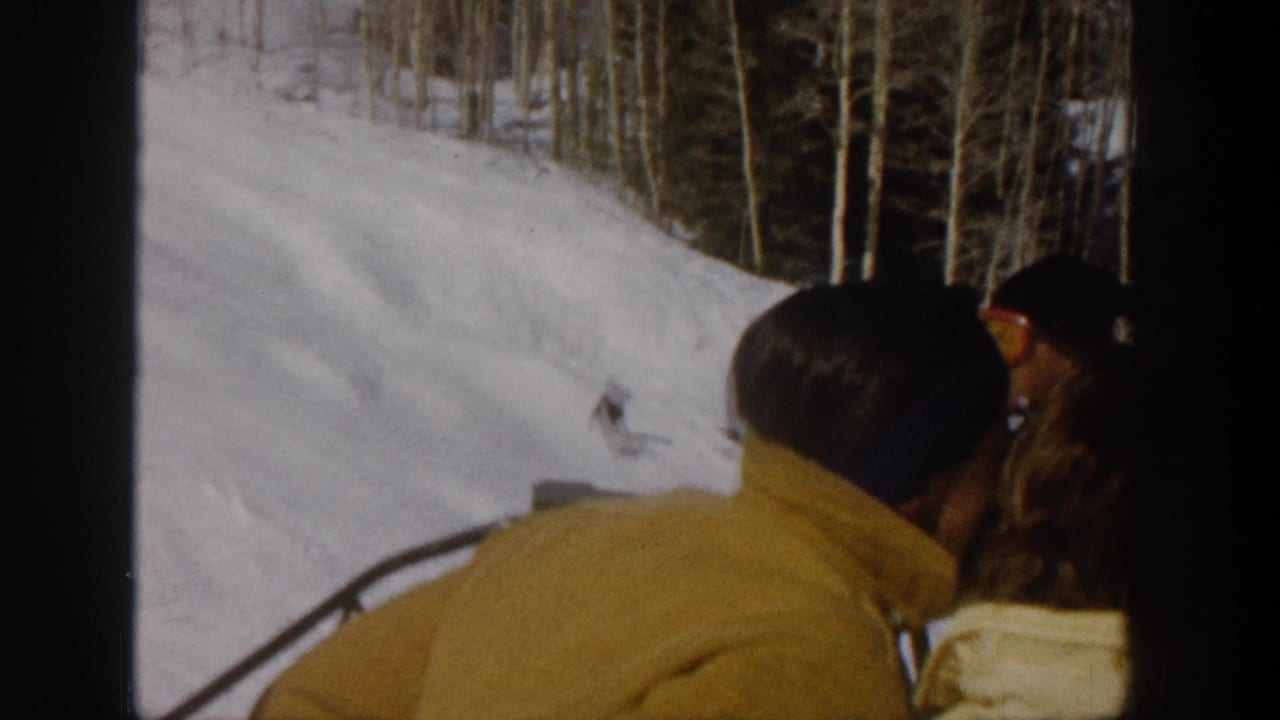 1961 Aspen home-movie frame showing a man in a yellow jacket riding a ski lift beside snowy terrain and bare aspen trees.
