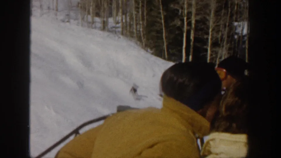 1961 Aspen home-movie frame showing a man in a yellow jacket riding a ski lift beside snowy terrain and bare aspen trees.