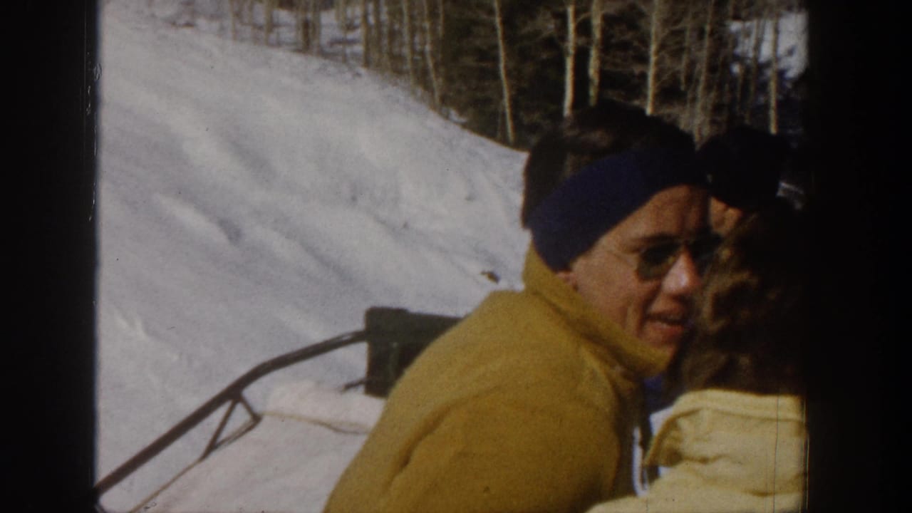 1961 Aspen frame of a ski-lift passenger in a yellow jacket and blue headband looking toward the camera with snowy slope behind.
