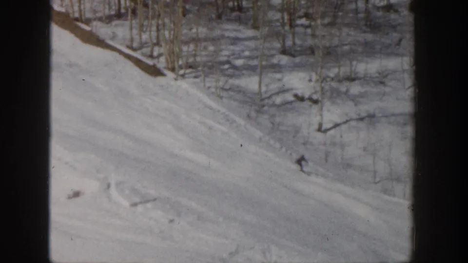 1961 Aspen home-movie frame with a small skier descending a snowy wooded slope, seen from a moving lift or elevated viewpoint.