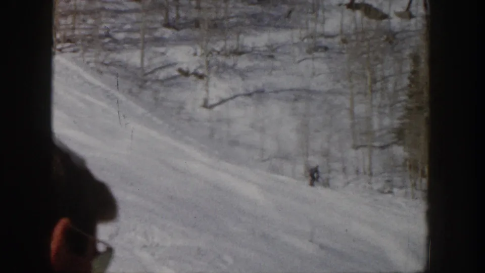 1961 Aspen frame showing a distant skier on a snow-covered slope while a nearby lift passenger is partly visible at the bottom edge.