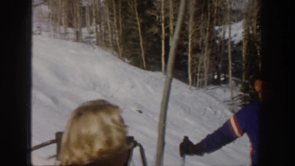 1961 Aspen 8mm frame showing a blonde woman in sunglasses in the foreground with skiers and trees on a snowy slope.