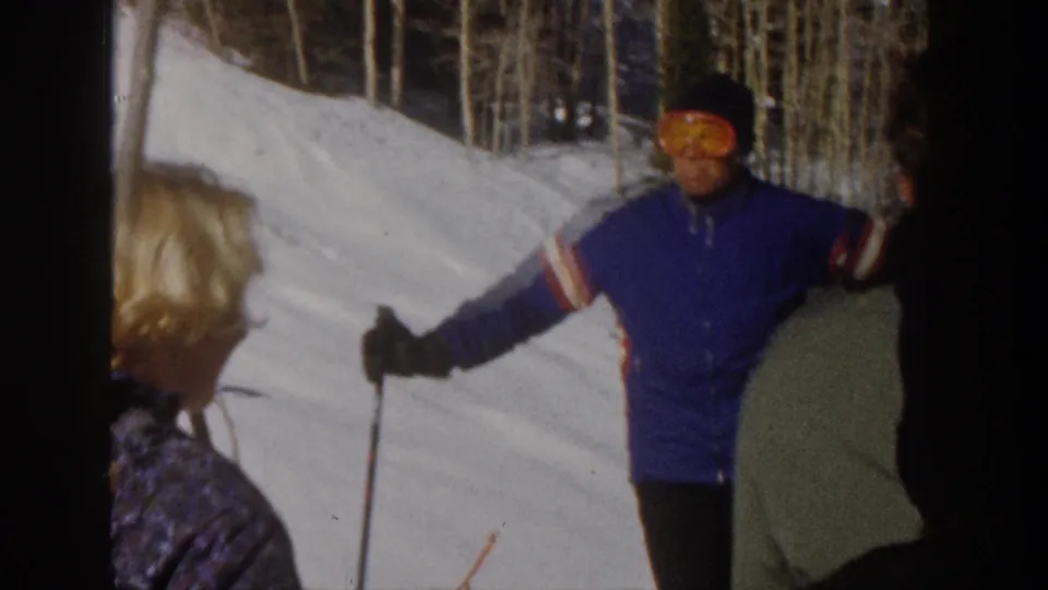 1961 Aspen 8mm frame of a man in a blue ski jacket and orange goggles gesturing toward the camera beside other skiers.