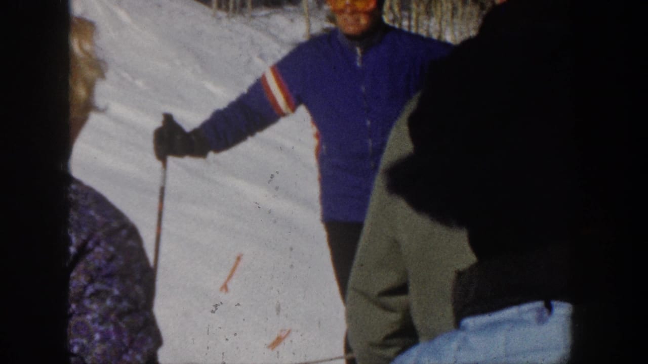 1961 Aspen home-movie frame showing skiers gathered on a snow-covered slope, with one person in a blue jacket and orange goggles holding poles.