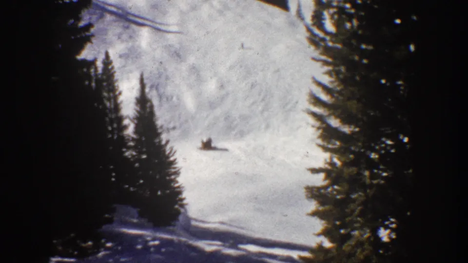 Wide 1961 Aspen ski frame with a small distant skier crossing a sunlit snowy slope between dark evergreen trees.