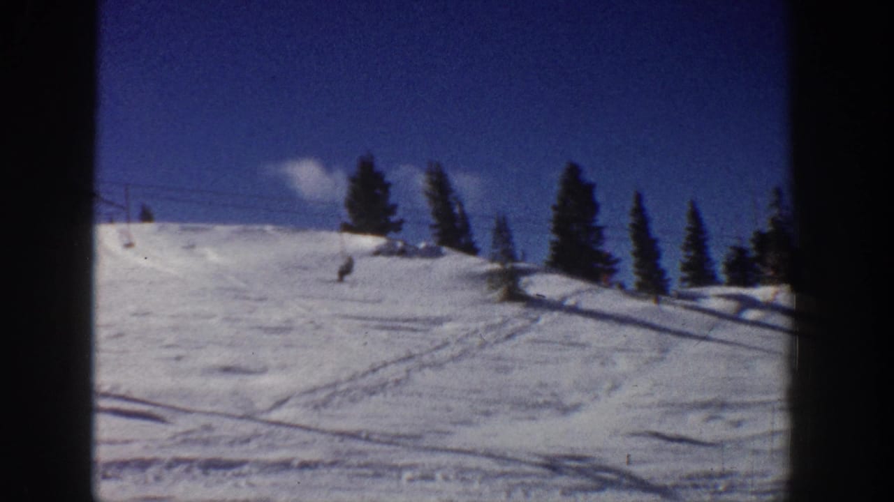 1961 Aspen ski slope with a distant skier, evergreen trees, lift infrastructure, and bright snow under blue sky.