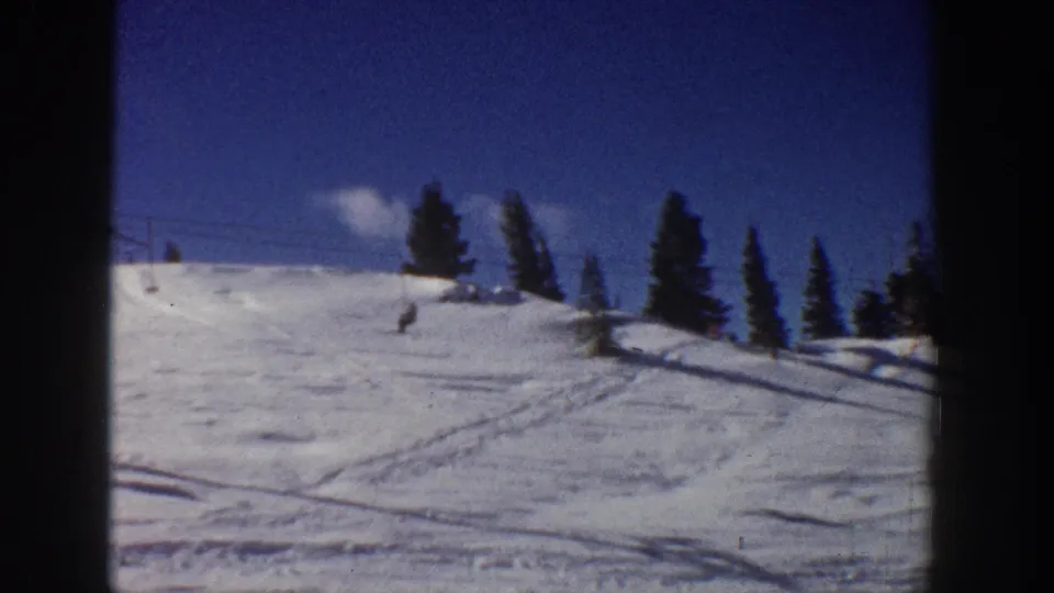 1961 Aspen ski slope with a distant skier, evergreen trees, lift infrastructure, and bright snow under blue sky.