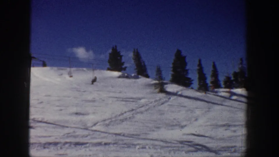 Wide 1961 Aspen ski-slope view with tracks in the snow, evergreen trees, and distant skiers or lift users.