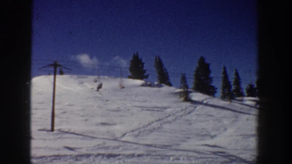 1961 Aspen winter frame showing a lift pole on a snowy ski slope with distant skiers and evergreen trees.