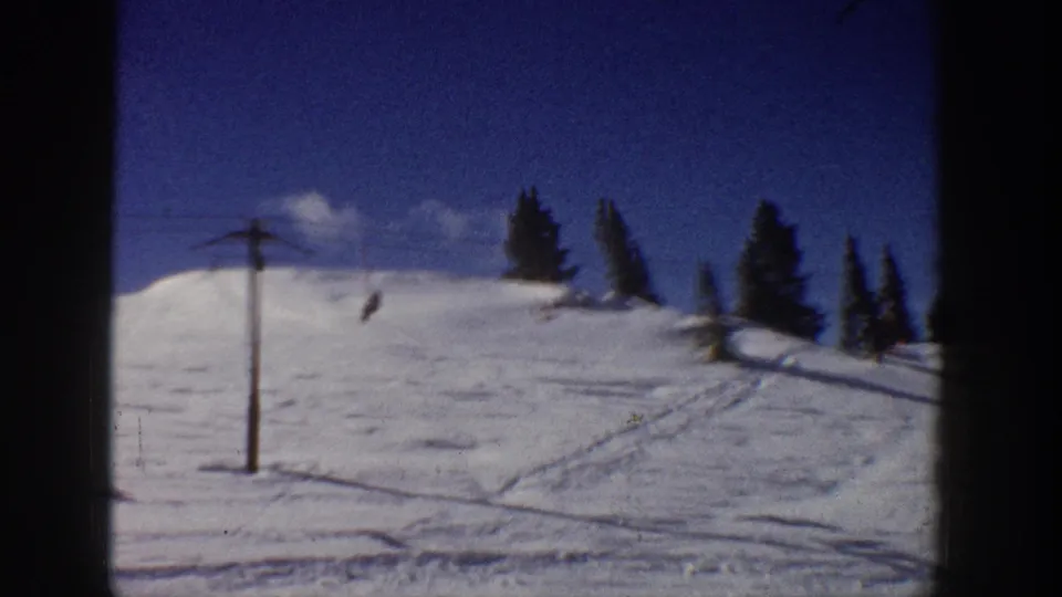 1961 Aspen home-movie frame showing a snowy ski slope, ski-lift pole and cable, evergreens, and tracks under a deep blue sky.