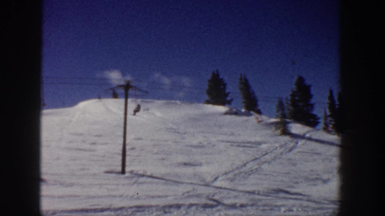 1961 Aspen frame of a snow-covered ski hill with lift equipment, trees, ski tracks, and small distant skier shapes.