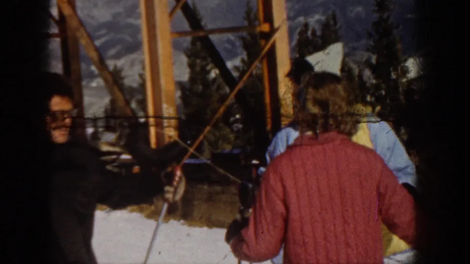 1961 Aspen lift-station frame showing adult skiers in jackets near wooden supports, with snow and mountain scenery in the background.