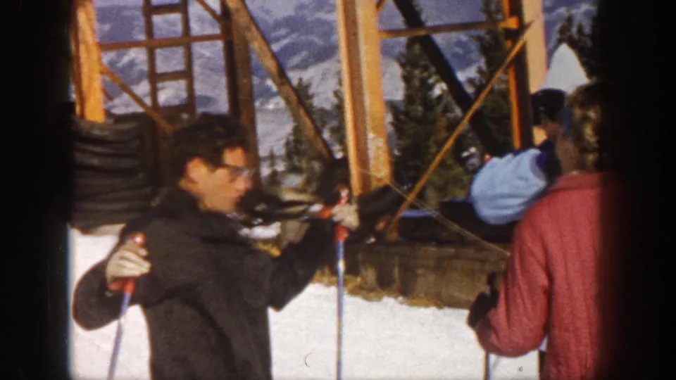 1961 Aspen home-movie frame of adult skiers at a lift station, including a man with glasses holding ski poles beside others in winter clothing.
