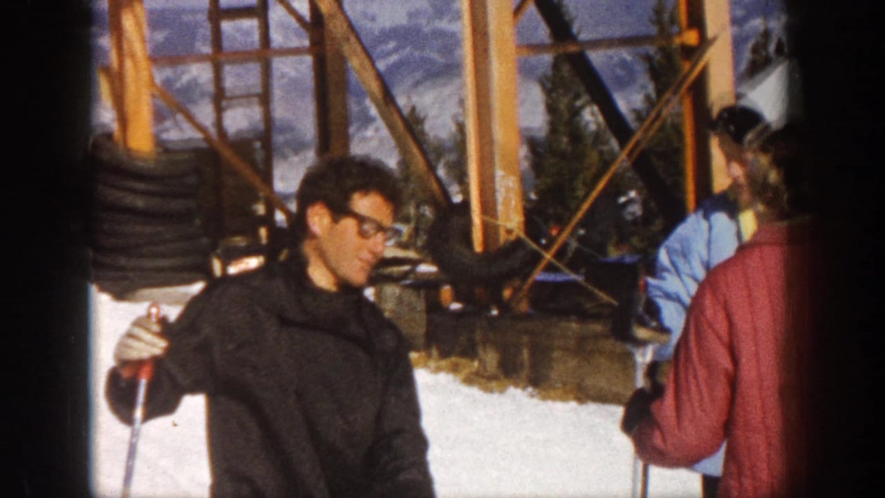 In Aspen, Colorado, 1961, a man in glasses holds ski poles beside a woman in red and a person in blue near snowy wooden lift structures.