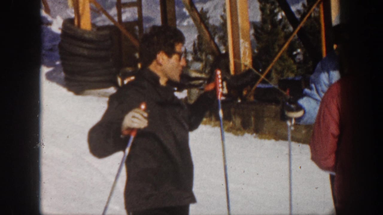 In the 1961 Aspen ski-resort scene, the man in a dark jacket gestures with poles while facing nearby skiers on snow.