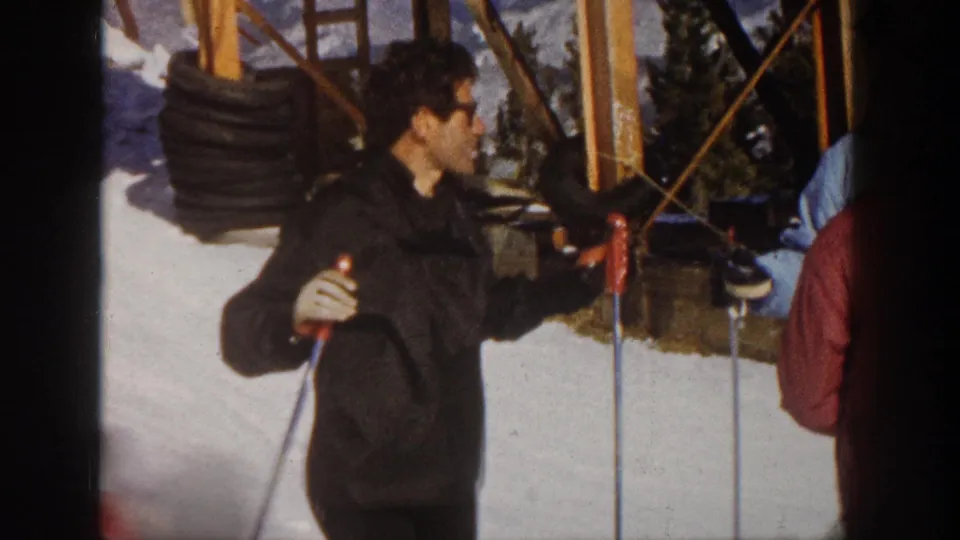 A candid 1961 Aspen frame shows a man with glasses holding ski poles as he speaks near two other skiers by the lift area.