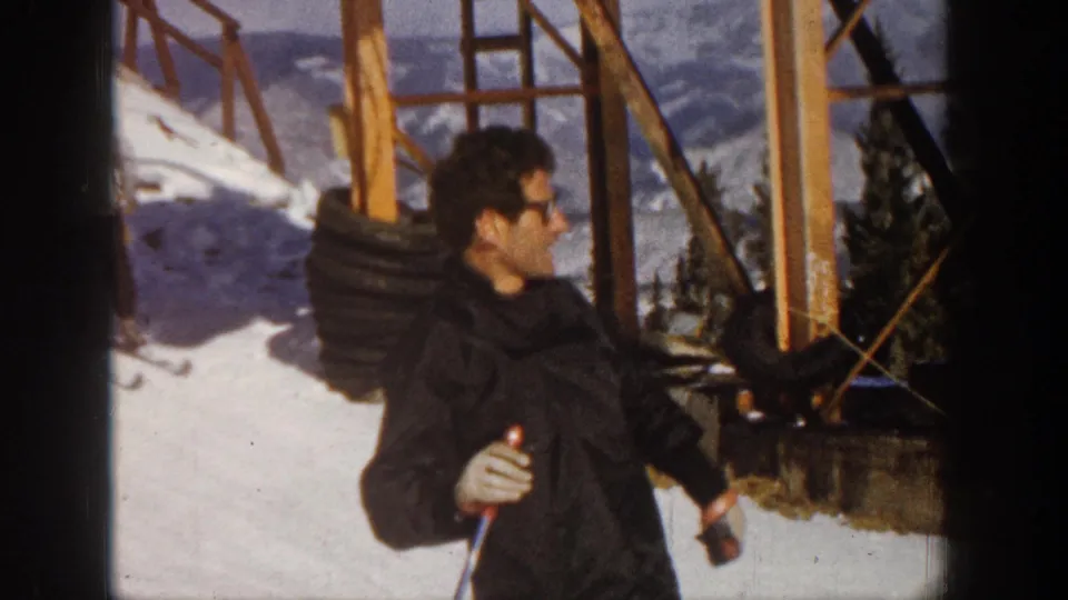 At Aspen in 1961, the skier in glasses looks aside with poles in hand, framed by snow, lift supports, evergreens, and distant mountains.
