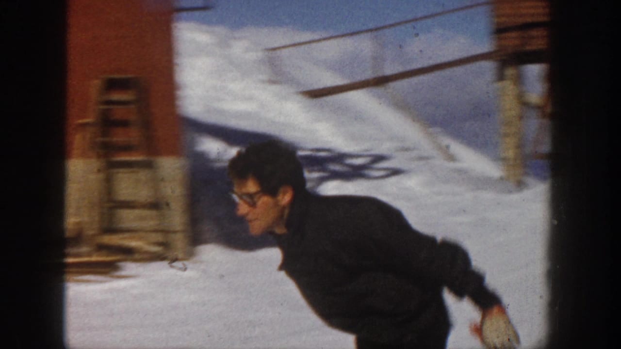 In Aspen, Colorado, 1961, a man in dark ski clothing and glasses leans forward while skiing past the camera on bright snow.