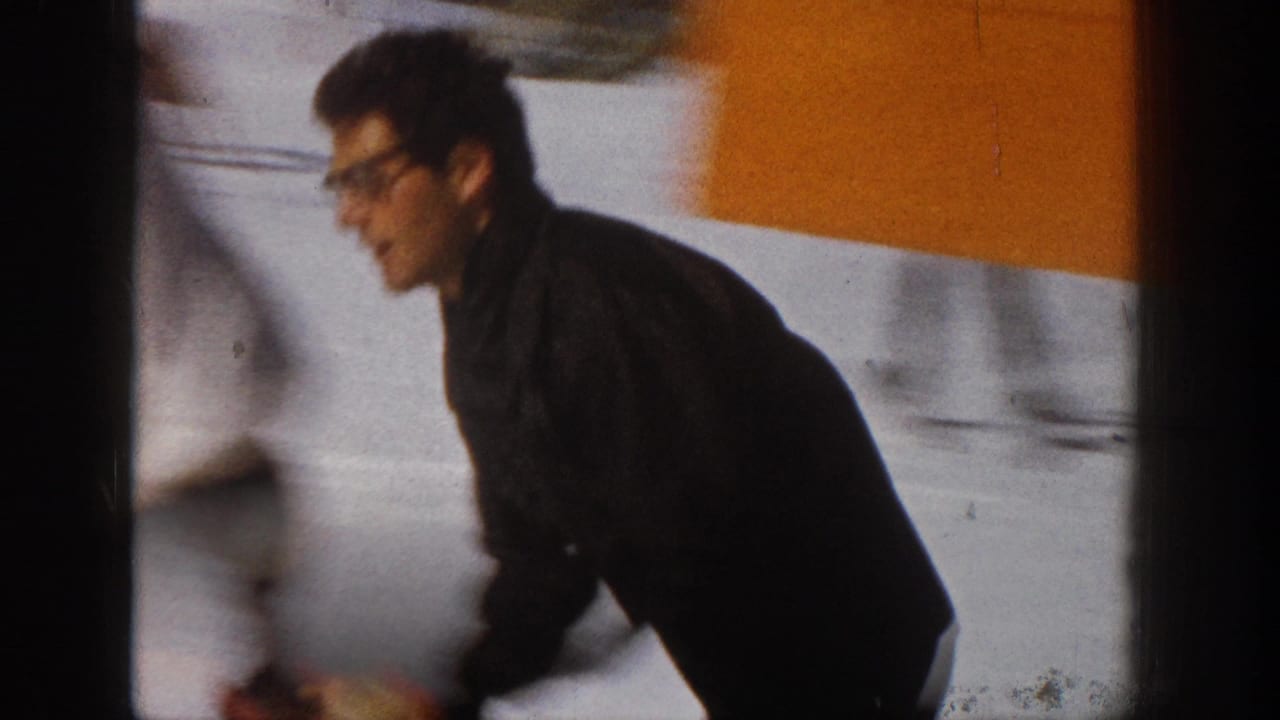 Man in dark ski clothing passes in profile on snow with an orange-and-white wall behind him.