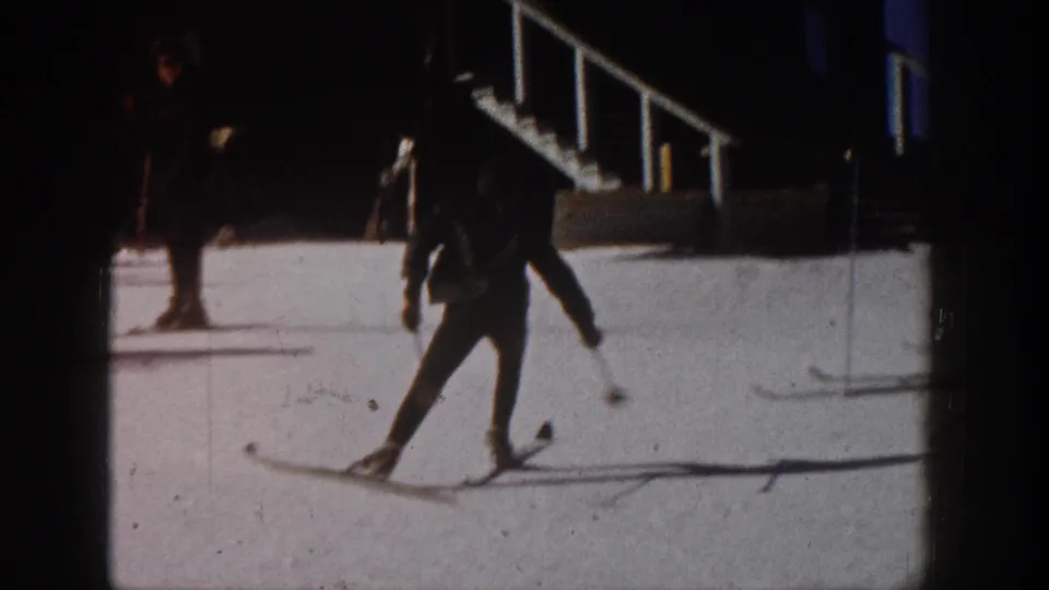 The adult skier glides across snow in a wider Aspen, Colorado, 1961 home-movie frame with railings and strong shadows.