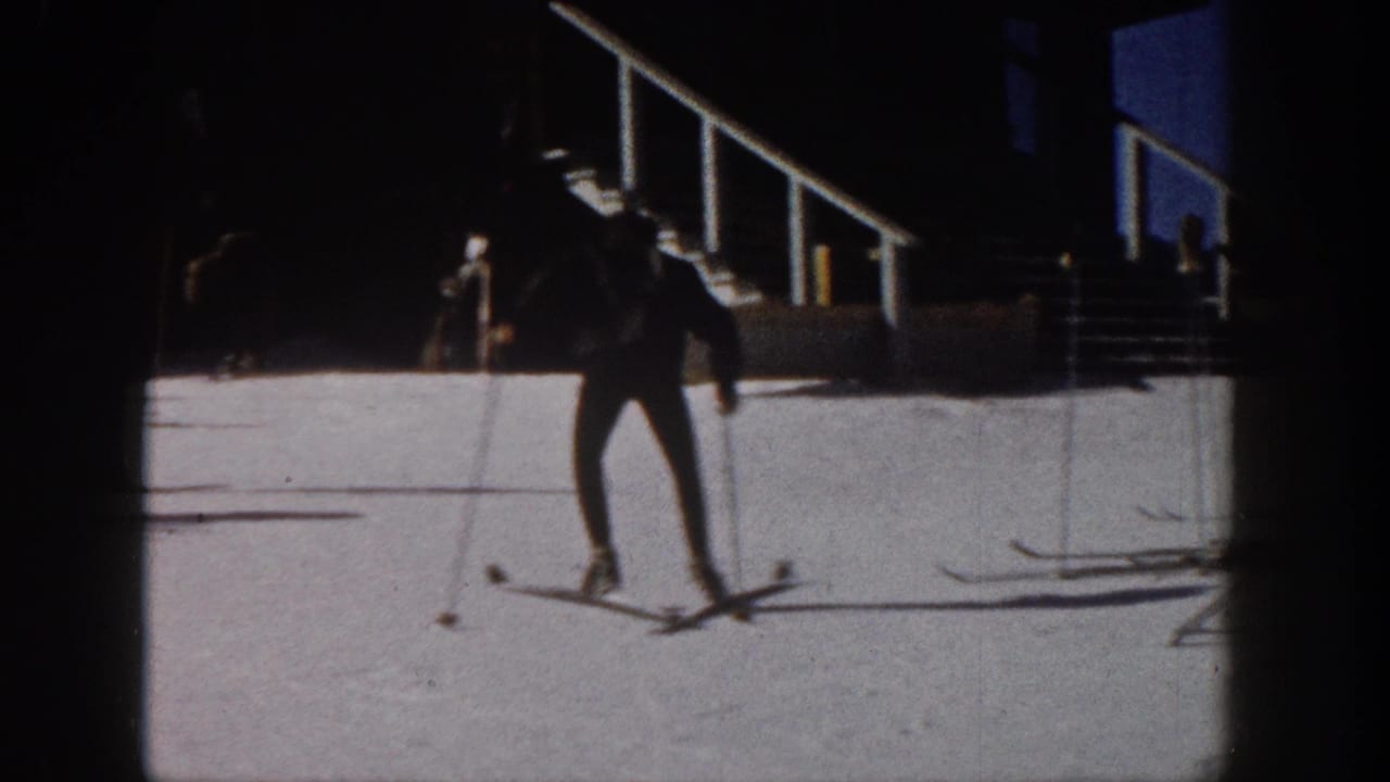 1961 Aspen home-movie frame showing a skier in dark winter gear on snow beside resort-side stairs and shadowed architecture.