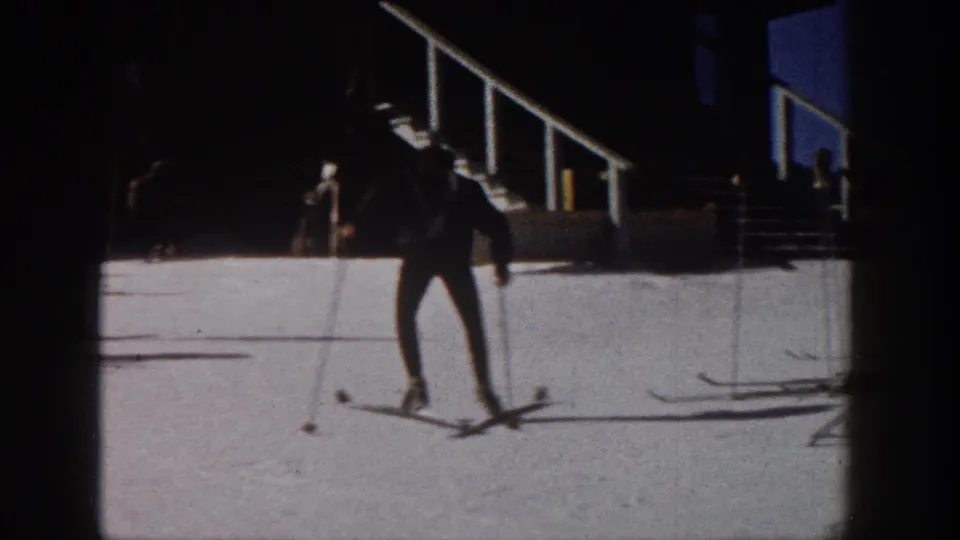 1961 Aspen home-movie frame showing a skier in dark winter gear on snow beside resort-side stairs and shadowed architecture.
