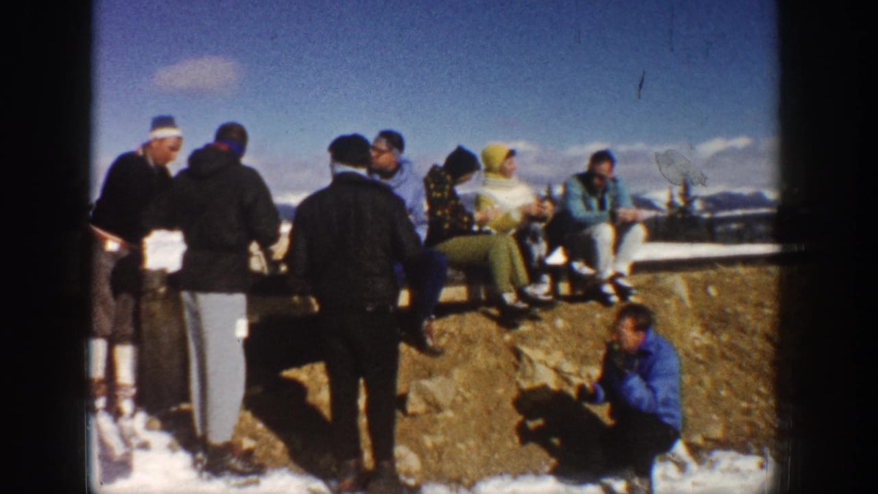 Bundled adults gather on rocky ground with snow patches and mountains in the distance.