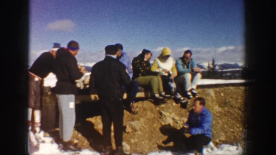 Friends in 1960s winter attire sit and stand at an Aspen overlook, with rocky ground, snow patches, and distant mountains behind them.