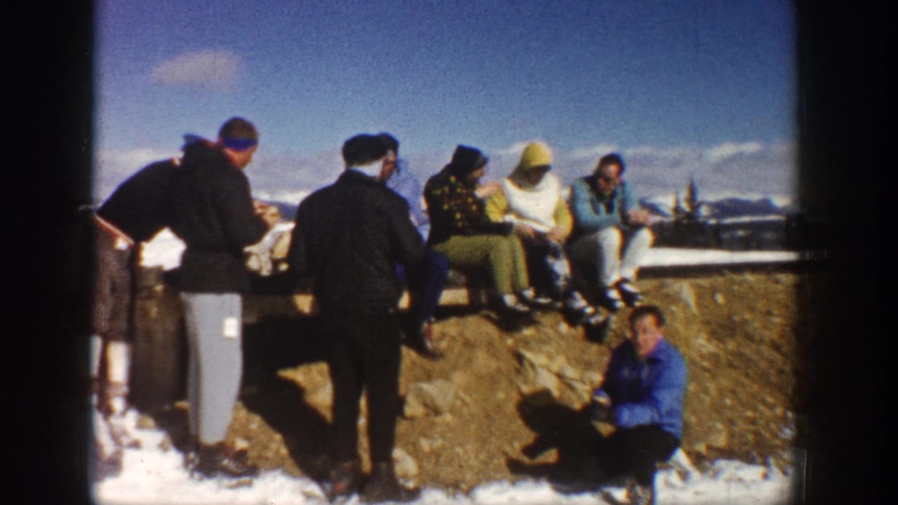 In Aspen, Colorado in 1961, adults in winter clothing sit and stand near a snowy ledge with distant mountains behind them.