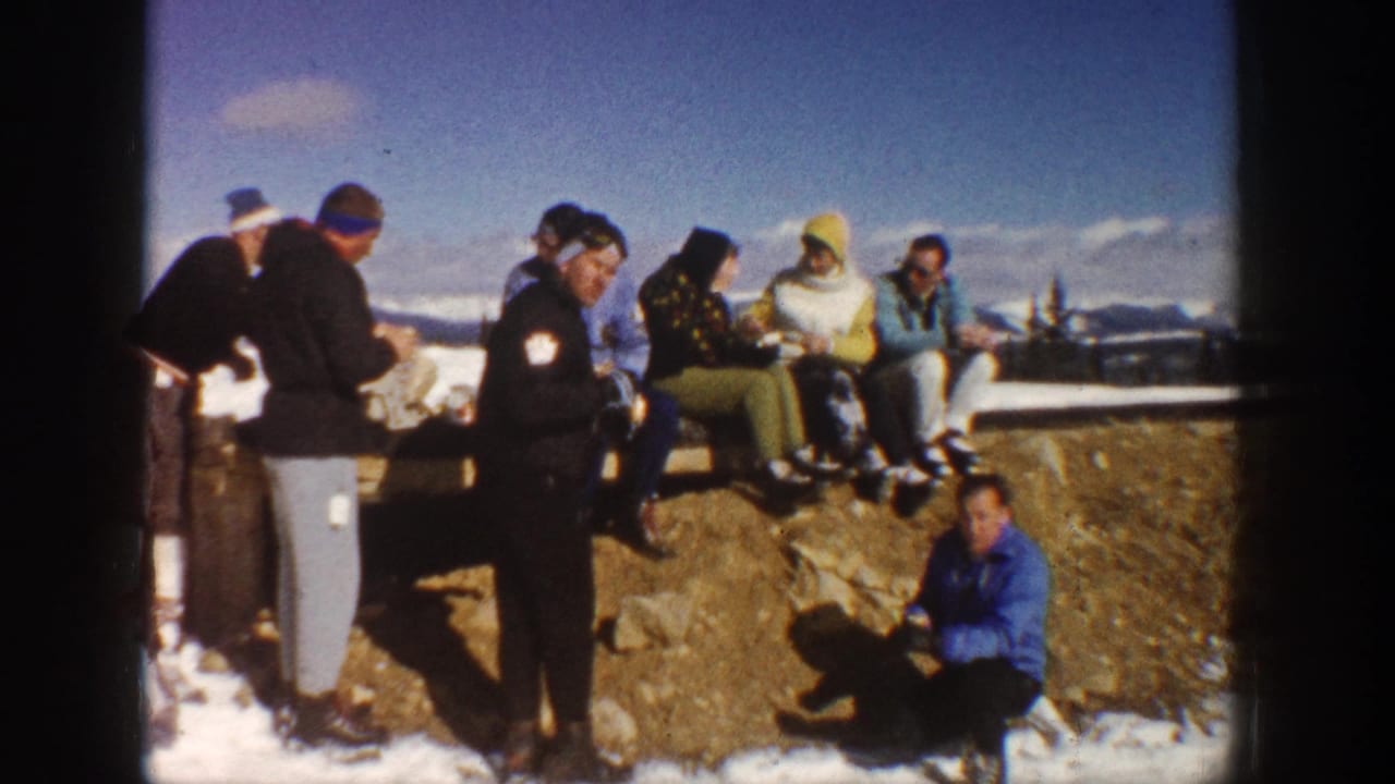 Adults in winter clothing sit and stand by a snowy ledge with mountains in the distance.