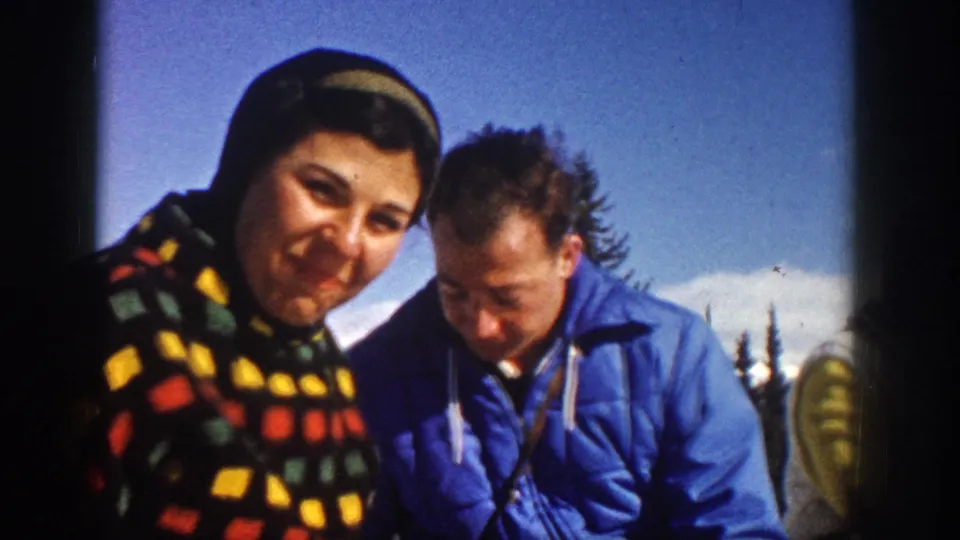 In the Aspen 1961 home movie, two friends sit close together in winter clothing, with evergreen trees and snow visible in the background.