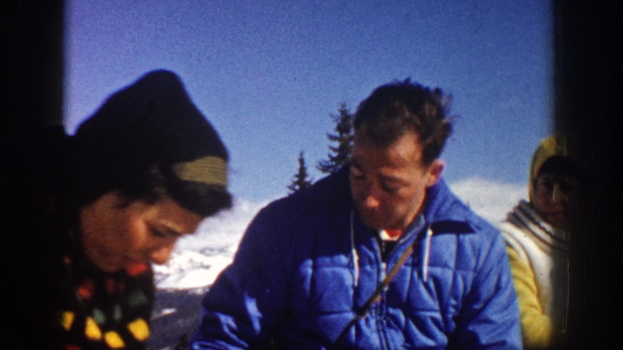 Three adults in winter clothing sit outdoors with snow and trees behind them.